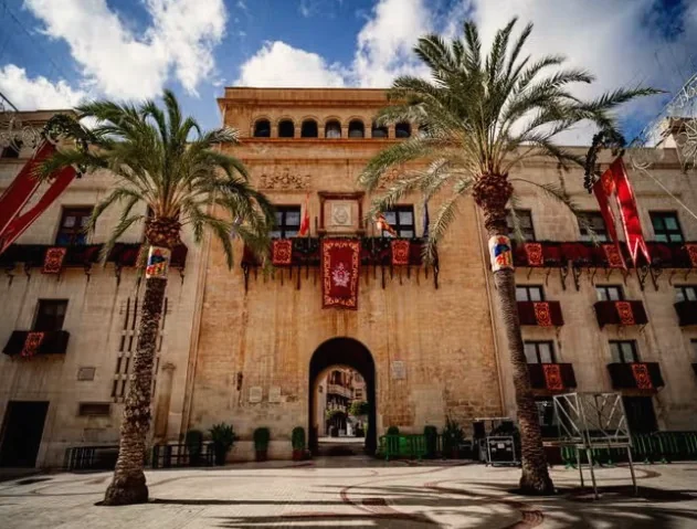 En bygning i sandstein med palmer og røde faner i gamlebyen i Elche